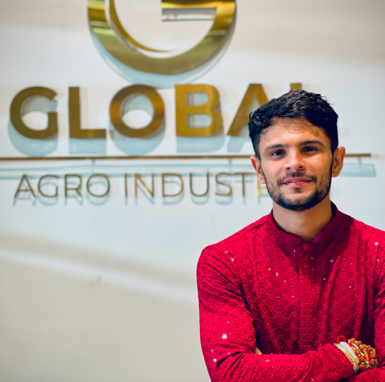 Manish Balyan wearing a bright red kurta, standing in front of the wall with global agro logo.