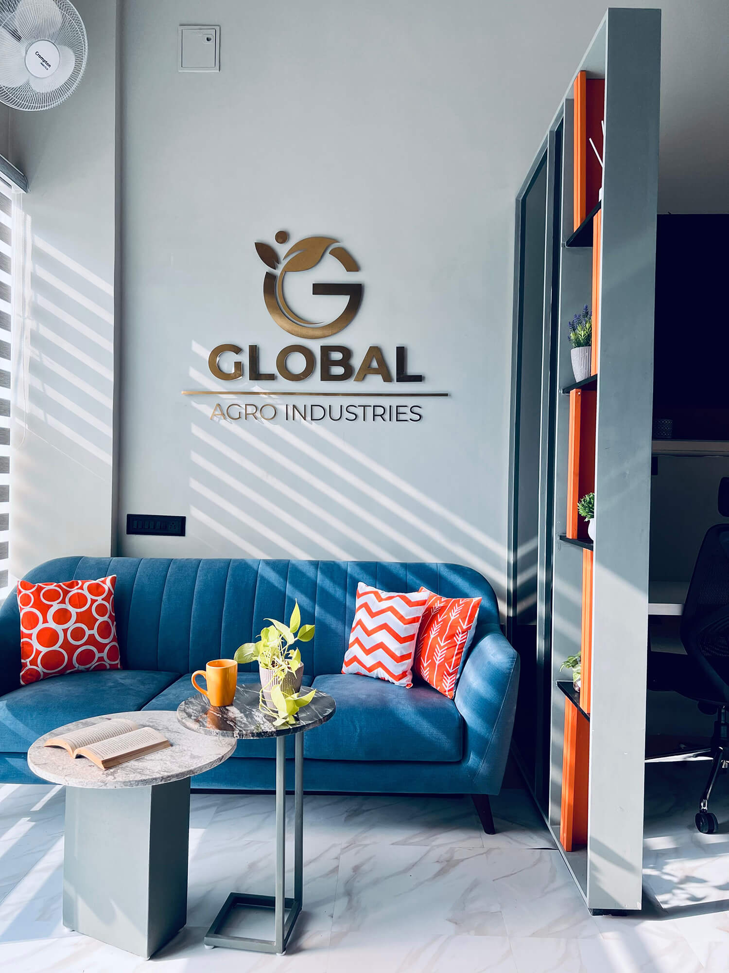 Office living room with a blue couch, wooden table, and a ceiling fan. Text on the wall says “Global Agro Industries”.