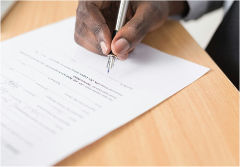 Hand of a dark skinned person holding a fountain pen over a document placed on a wooden surface.